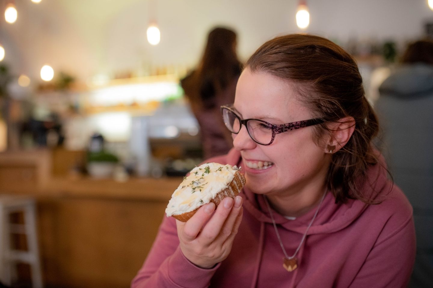 Mlada žena sjedi u kafiću, smije se dok razgovara s prijateljicom i u ruci drži krišku kiselog kruha s namazom od pečenog češnjaka